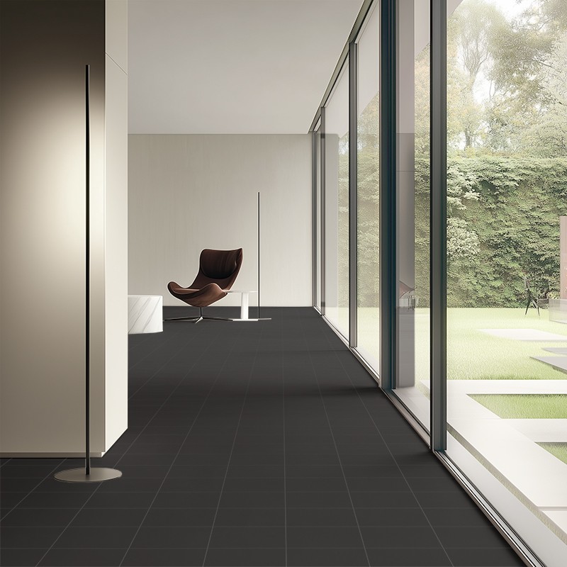 Piso de azulejos pretos foscos (foco) em corredor amplo; parede de vidro panorâmica para jardim gramado, poltrona marrom minimalista e luminárias finas.