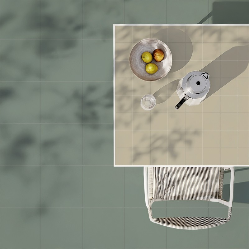 Vierkante lichtbeige tegel met espresso-koffiepot, klein glas en kom met drie appels; smalle witte rand; bovenaanzicht, stoelrand en groene tegelvloer met gefilterde schaduwen op de achtergrond.