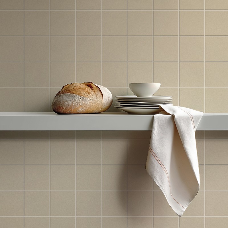 Azulejo beige cuadrado en pared; repisa blanca con pan redondo, platos y cuenco apilados y paño colgante, ambiente de cocina minimalista y luminoso.