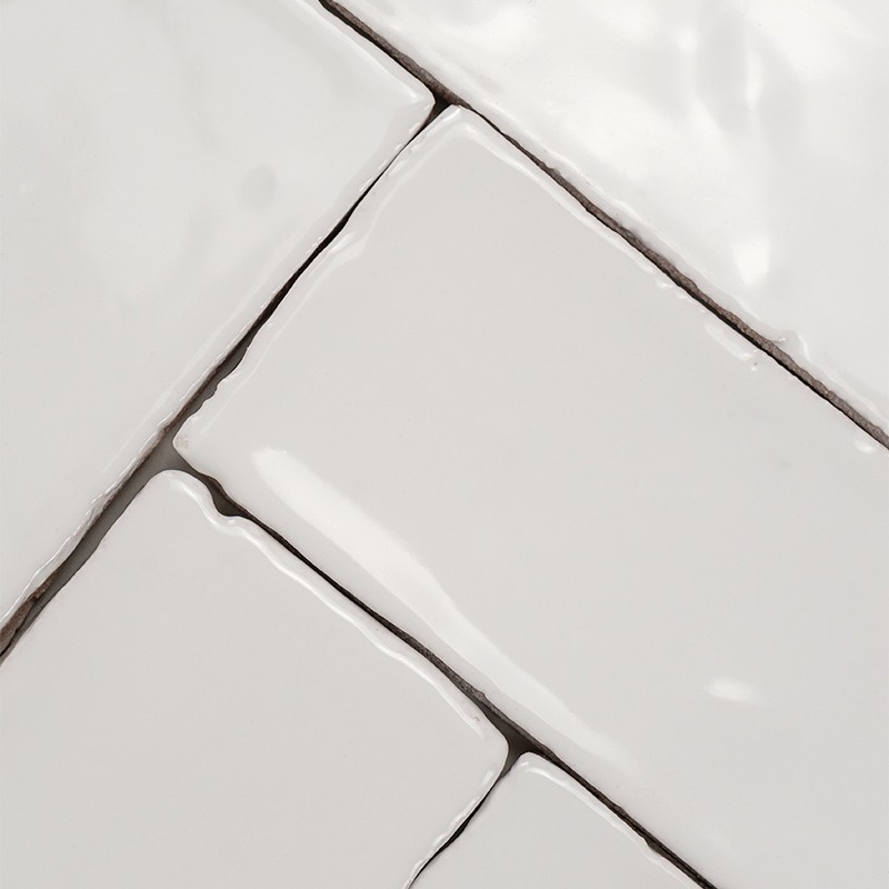 Witte geglazuurde keramische tegels met licht glooiende randen, zichtbare donkere voegen en glanzende reflecties; diagonaal gerangschikt, subtiel oneffen oppervlak.