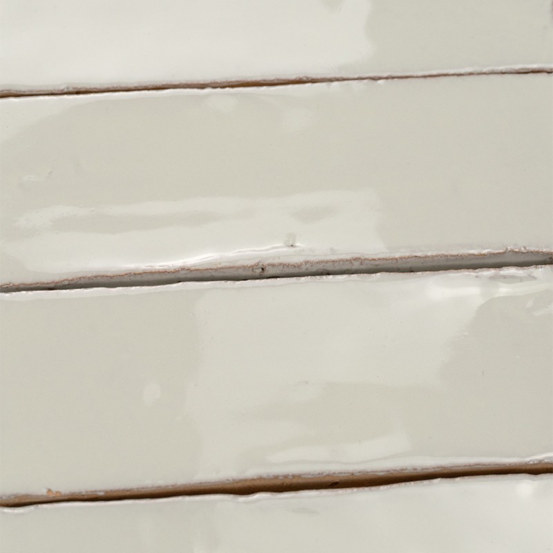 Azulejos cerâmicos retangulares brilhantes, cor creme; alinhados horizontalmente com rejuntes finos marrons, pequenas irregularidades na película de esmalte e reflexos.
