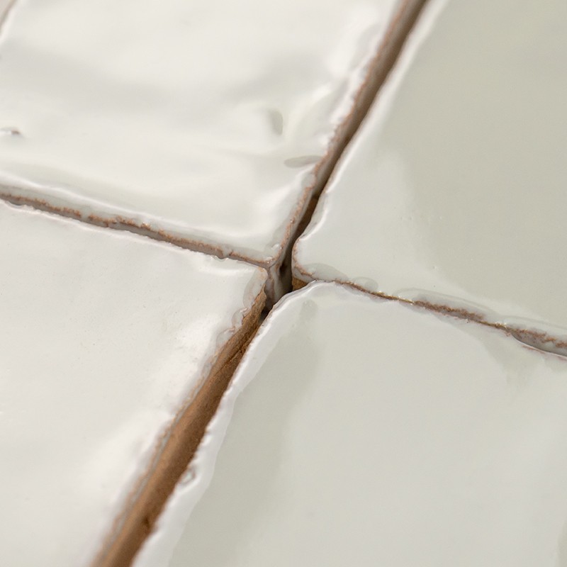 White glazed ceramic tile with beige grout joints, cross joint showing small imperfections and shine, slightly irregular texture and rounded edges.