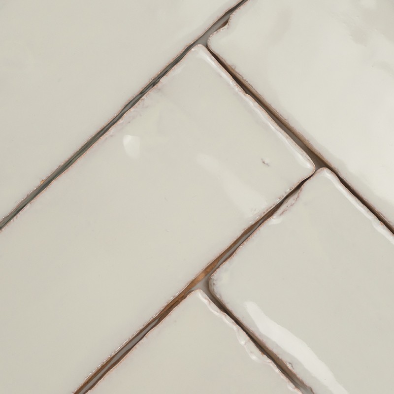 Azulejo cerâmico retangular branco com esmalte brilhante, cantos e juntas levemente irregulares e sujeira marrom nas fendas; close-up de revestimento de parede.