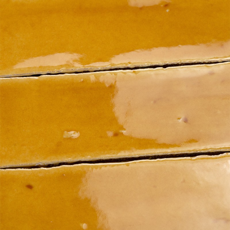 Carrelage céramique jaune-ocre brillant, jointures horizontales foncées, surfaces émaillées avec reflets, variations de teinte et petites imperfections visibles.