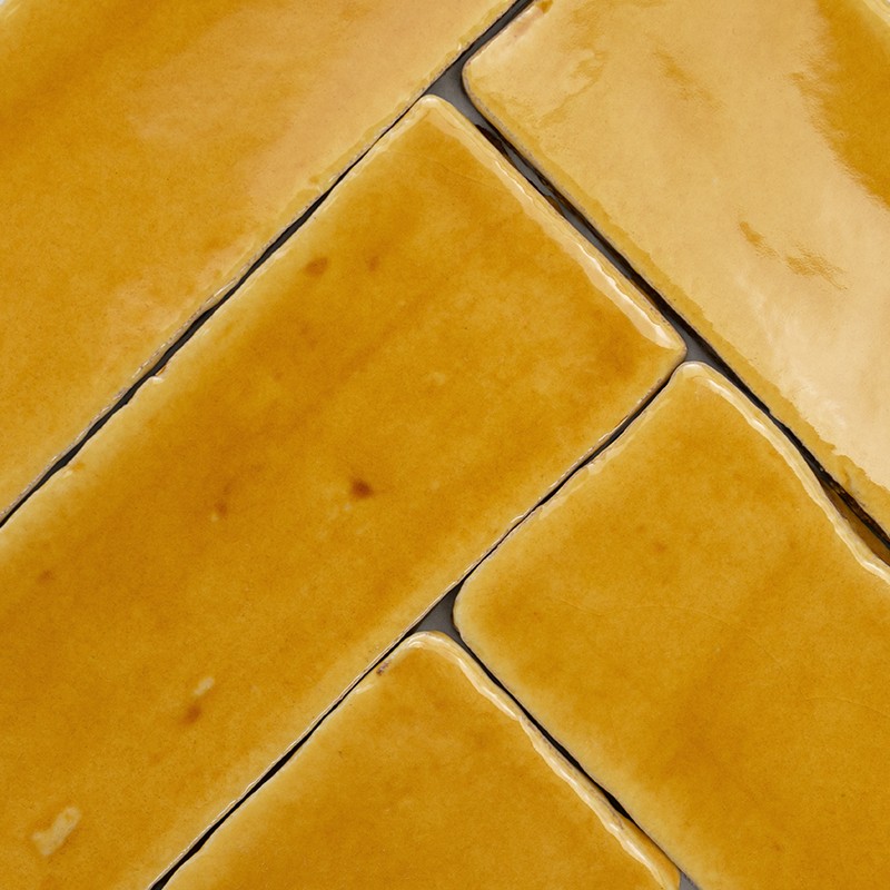 Bright yellow rectangular tiles in a herringbone pattern, pronounced dark joints; glazed surface with small spots and wear, close-up view.