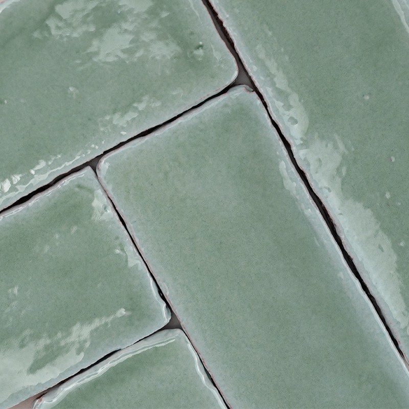Azulejo esmaltado verde pálido y rectangular; juntas oscuras hundidas forman patrón entrelazado, superficie brillante con reflejos y pequeñas imperfecciones.