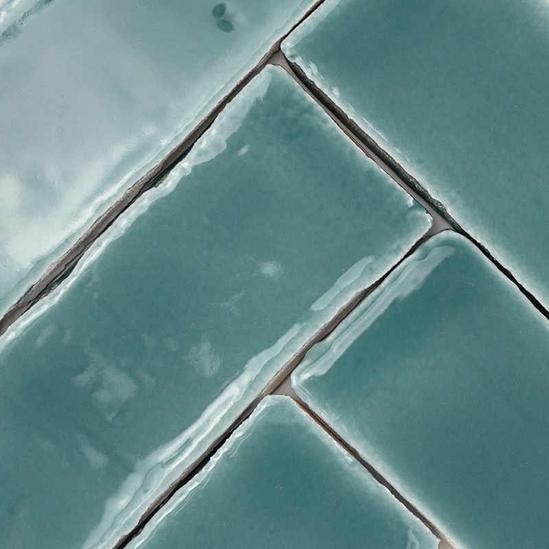 Azulejo cerâmico retangular verde-azulado brilhante com esmalte irregular; juntas escuras e desgastadas entre peças formando padrão diagonal.