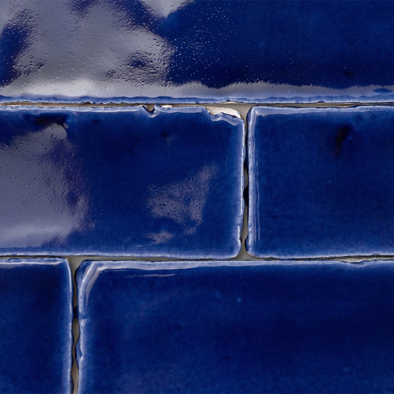 Azulejos cerâmicos retangulares azul profundo, superfície vidrada com reflexos e pequenas irregularidades; rejuntes claros entre peças, iluminação suave realçando brilho.