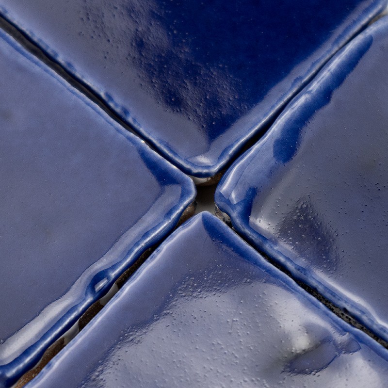 Glänzende dunkelblaue Keramikfliese mit zentraler Kreuzfuge; vier leicht gewölbte Quadranten, helle Glanzreflexe, feine Risskanten und dunkle Fugenmasse sichtbar.