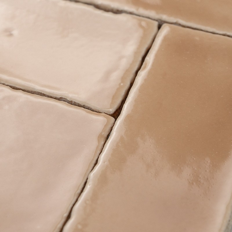 Beige-braune, glänzende Keramikfliesen mit unregelmäßigen Kanten und sichtbaren hellen Fugen; Nahaufnahme zeigt mehrere nebeneinander liegende, leicht unebene Oberflächen.