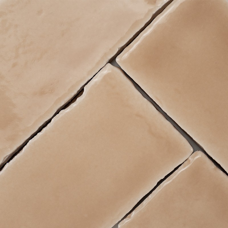 Beige keramische tegel met licht geglazuurde, afgeronde, onregelmatige randen; donkere voeglijnen in diagonaal patroon; subtiele kleurvariaties en lichte oppervlakteglans.