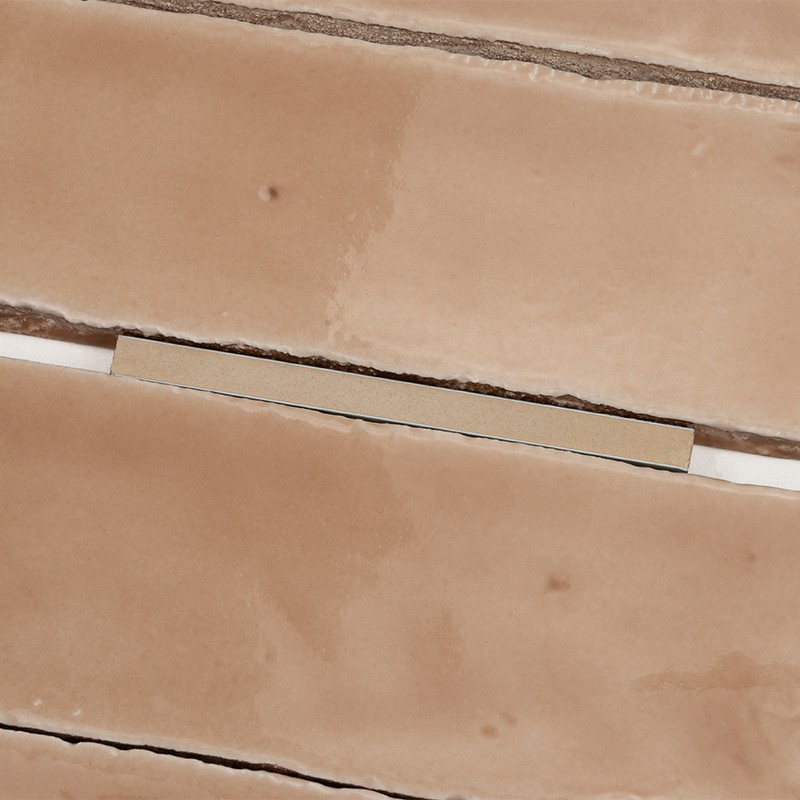 Azulejo rectangular esmaltado beige, brillo reflectante y pequeñas imperfecciones; dispuesto horizontalmente entre juntas estrechas de lechada oscura en un revestimiento continuo.