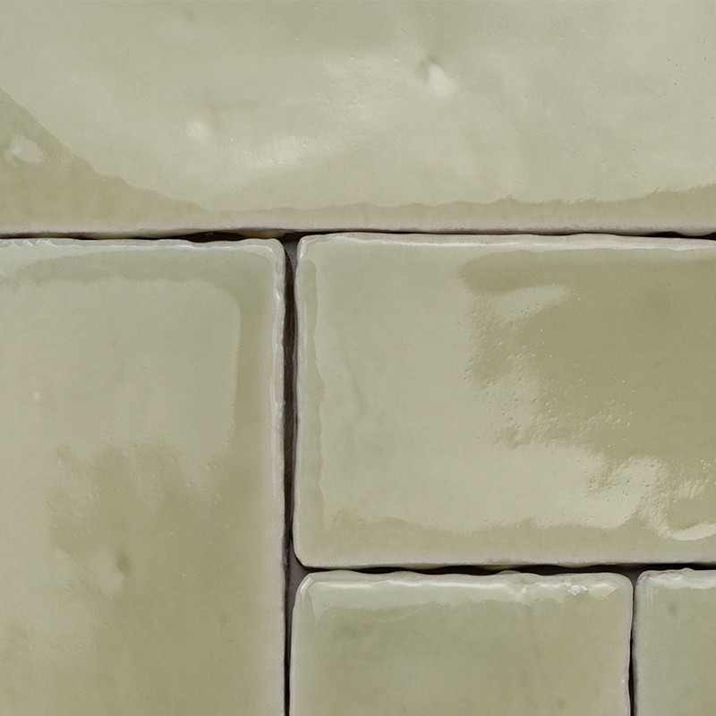 Rechteckige, beige-olivfarbene, glänzende Fliese mit sichtbaren dunklen Fugen und leicht unregelmäßiger Oberfläche; Nahaufnahme eines gefliesten Wand- oder Bodenbereichs.