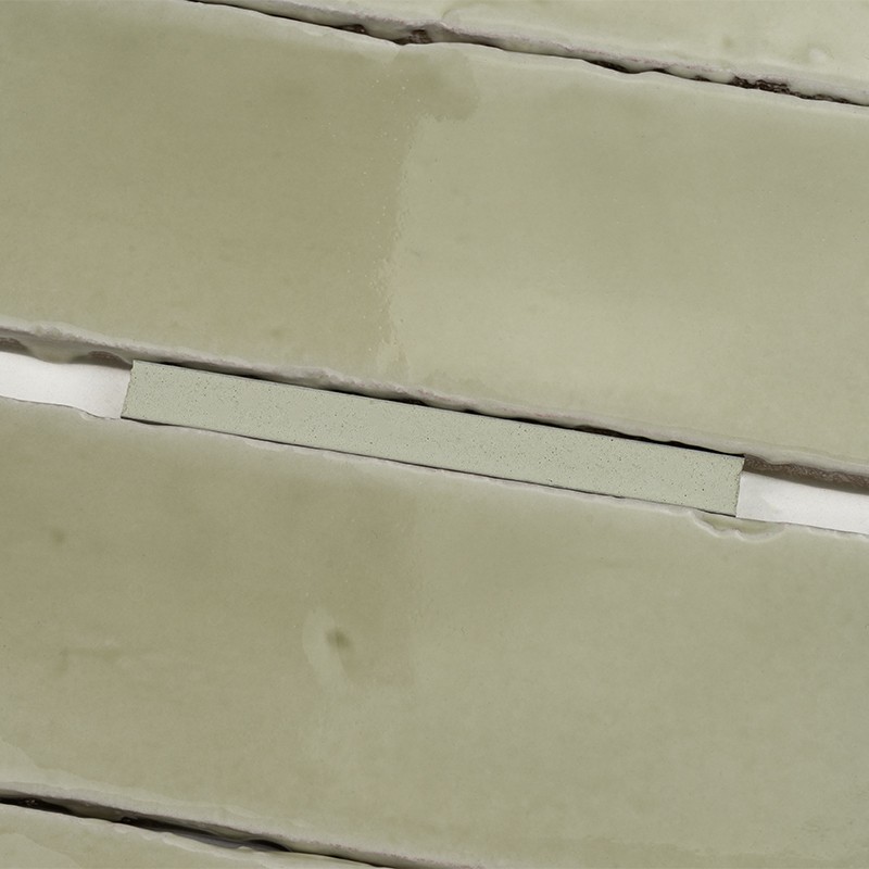 Azulejo rectangular esmaltado color verde pálido, borde irregular y ligeras imperfecciones; colocado horizontalmente en fila con juntas blancas y huecos estrechos entre azulejos.