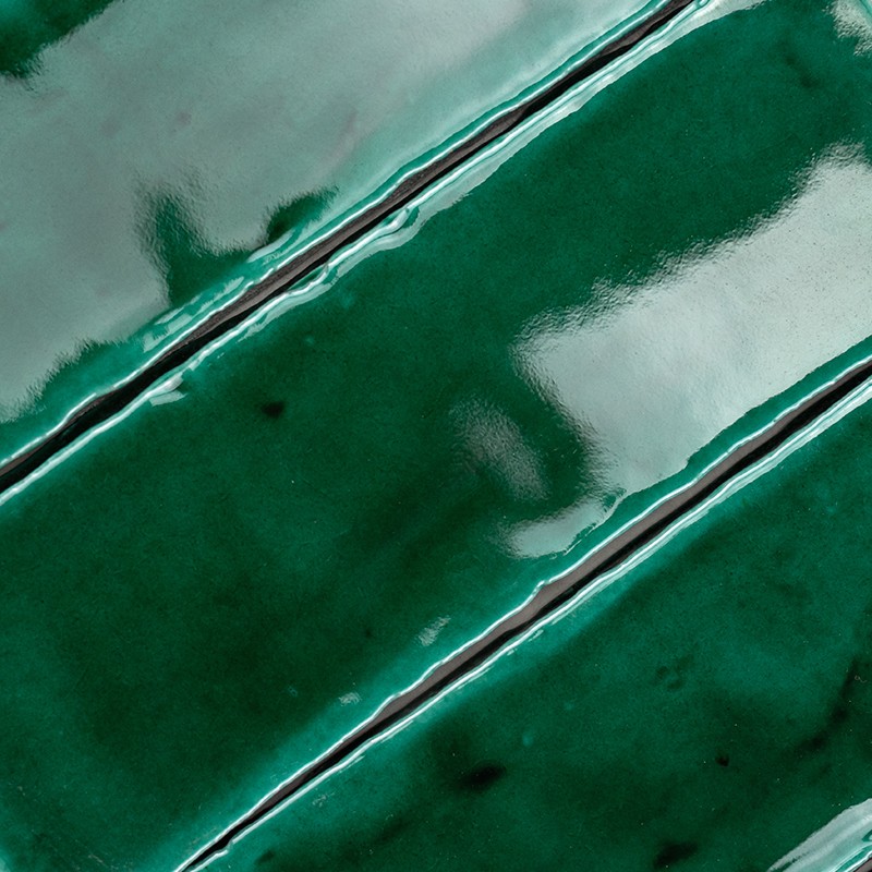 Azulejo esmaltado verde oscuro con tres ranuras diagonales claras, textura vidriada brillante y pequeñas manchas, bordeado por azulejos similares.