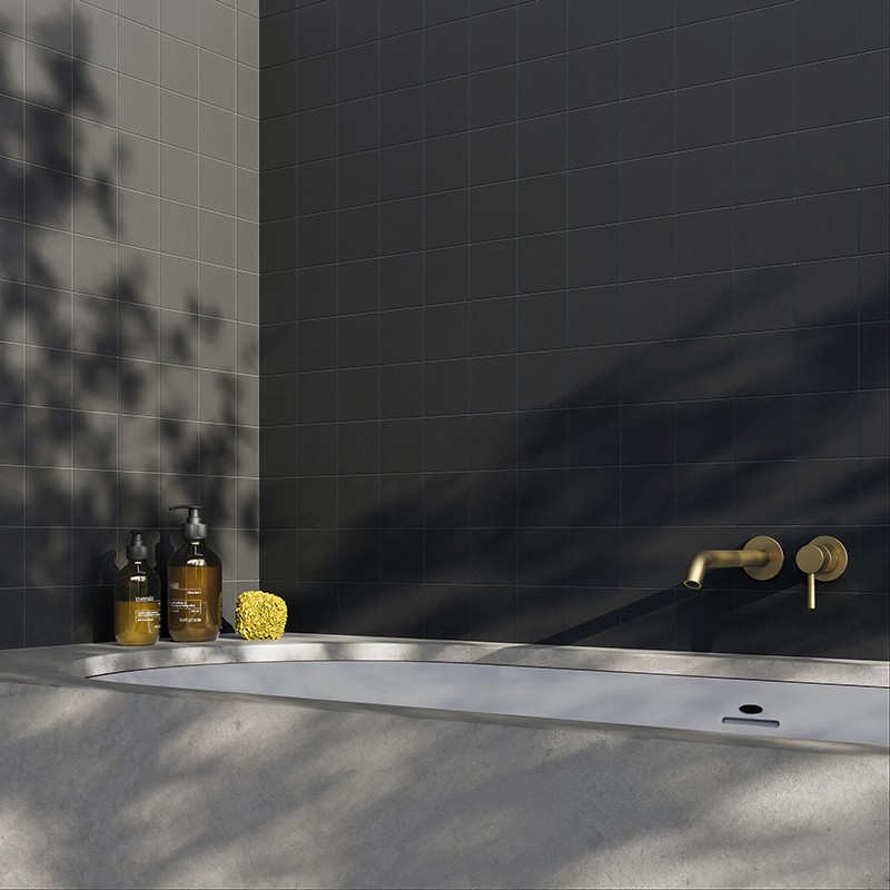Dunkle matte Wandfliesen über einer weißen Badewanne; goldfarbener Wandmischer, drei Pflegeflaschen und ein gelber Schwamm auf dem Wannenrand; diffuse Pflanzenschatten.
