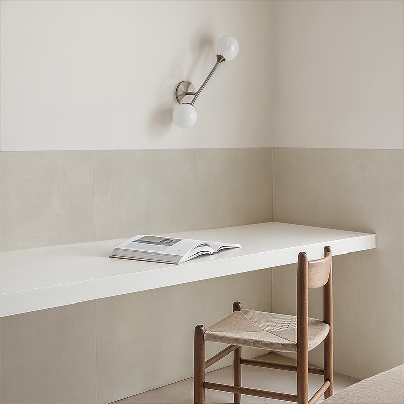 Azulejo beige liso en zócalo medio muro; entorno: escritorio flotante blanco con libro abierto, silla de madera con asiento trenzado y aplique de pared de dos esferas.