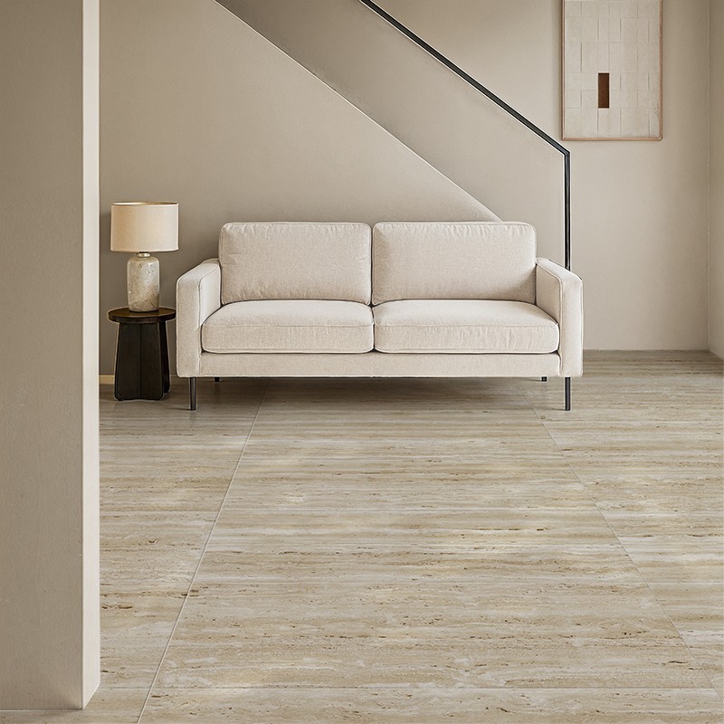Carrelage grand format imitation bois clair au premier plan; salon minimaliste aux tons neutres, canapé beige, lampe sur petite table sombre et mur angulaire beige en arrière-plan.