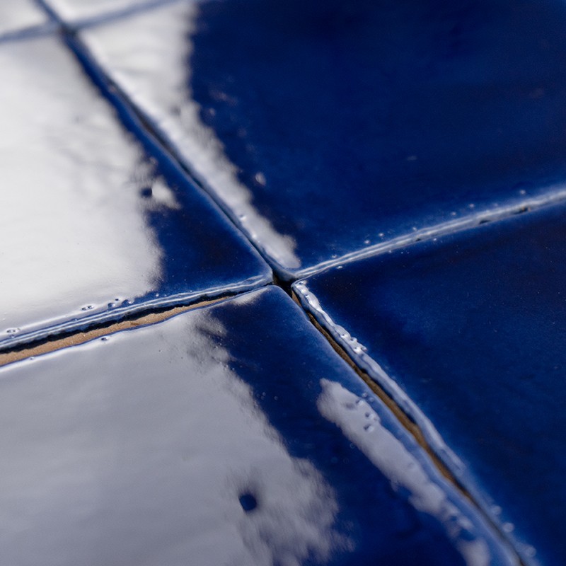 Dark blue ceramic tile, glazed shine with reflections; four-piece joint and visible seams, with small imperfections and relief in the foreground.