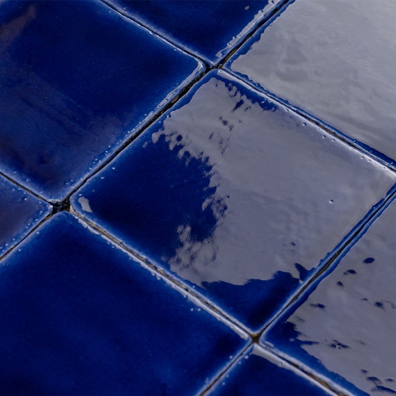 Intense blue square ceramic tile, glazed and reflective surface; narrow and dark joints, repeated arrangement forming a wall or floor covering.