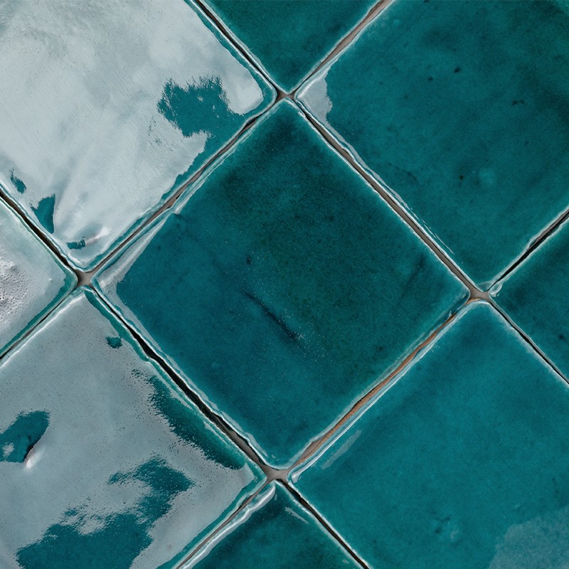 Bright and textured turquoise ceramic tile, square in shape and placed diagonally; surrounded by light joints that form a repeated pattern with slight reflections and variations in tone.