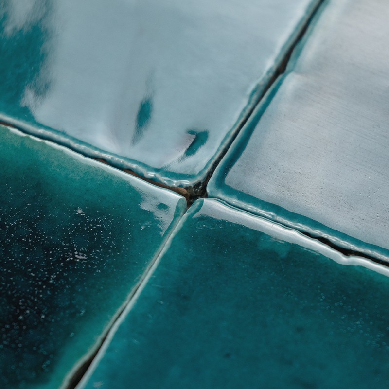 Blue-green glazed tile in the foreground; gray grout joints and wear, bright reflections and small bubbles, surrounded by adjacent tiles.