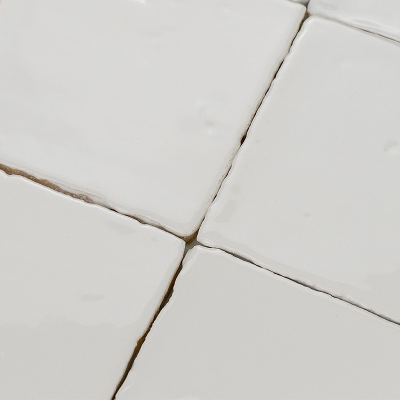 Azulejos cuadrados blancos esmaltados, juntas de lechada agrietada entre ellos; superficie plana con ligera textura e imperfecciones visibles bajo luz suave.