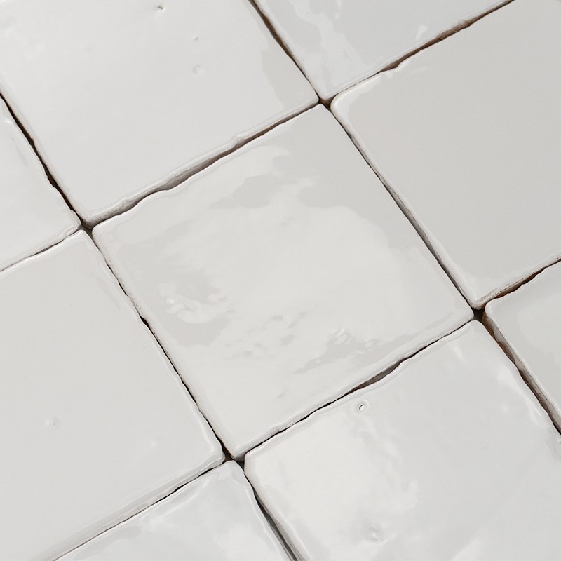 Witte, vierkante glanzende keramische tegels met licht geglazuurde, onregelmatige randen en smalle voegen, dicht tegen elkaar geplaatst onder diagonale hoek.