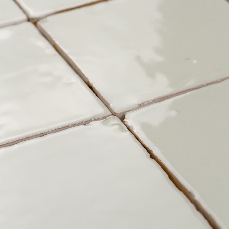 Geglazuurde witte vierkante tegels met zichtbare, licht verkleurde voegen; glanzend oppervlak met subtiele reflecties, enkele randen licht beschadigd en tegels dicht tegen elkaar gelegd.