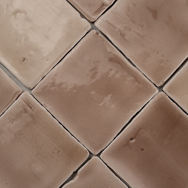 Azulejos cerámicos cuadrados dispuestos en rombo, superficie beige brillante con variaciones y pequeñas abolladuras; juntas oscuras visibles entre piezas sobre una superficie continua.