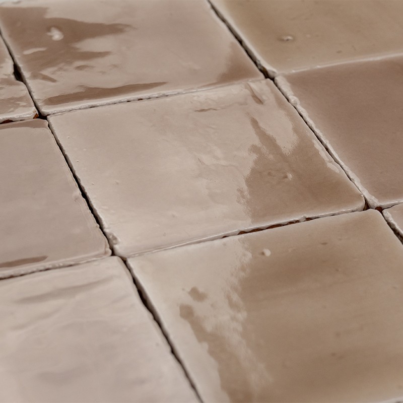 Glänzende beige quadratische Keramikfliesen, vermutlich Boden-/Wandbelag, in Nahaufnahme; sichtbare Fugen, unregelmäßige Glasur, Lichtreflexe und leichte Oberflächenunebenheiten.