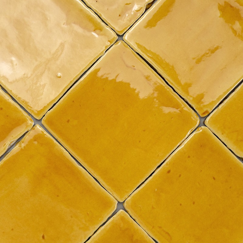 Gelbe, glänzende quadratische Fliese (diagonal platziert) mit dunklen Fugen und unregelmäßiger Glasur; angrenzende gleiche Fliesen bilden ein gekacheltes Feld.