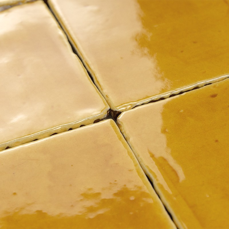 Azulejo amarillo vidriado, bordeado por lechada blanca agrietada; vista en detalle de cuatro piezas que confluyen en una esquina, refleja luz sobre superficie brillante.