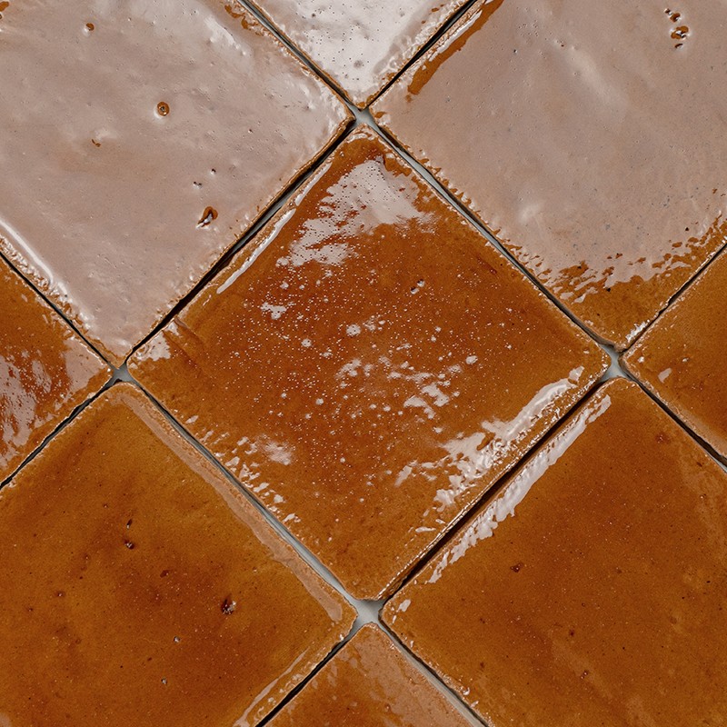 Azulejo cuadrado esmaltado color marrón anaranjado dispuesto en diagonal; juntas estrechas grises, brillo irregular con pequeñas burbujas y manchas de esmalte.