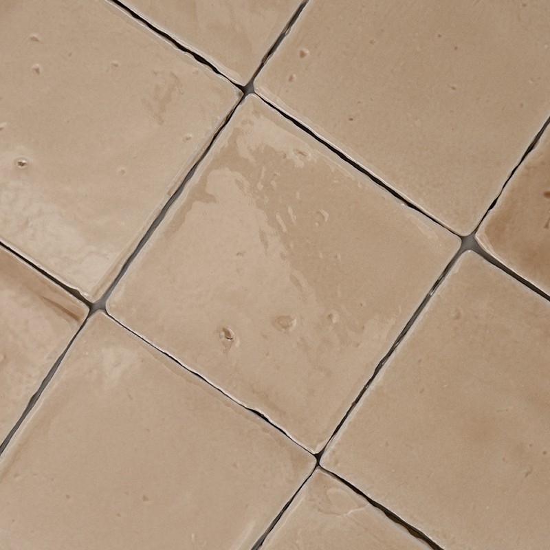 Een beige geglazuurde vierkante tegel met lichte onregelmatigheden, kleine putjes en glans; nauw met donkere voegen geplaatst in een raster op een vlakke wand of vloer.