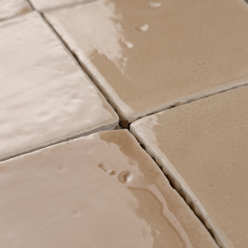 Geglazuurde beige vierkante tegels met zichtbare voegen; water parelt en vormt kleine plasjes bij de kruisende voegpunten, licht reflecterend.