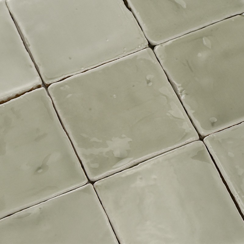 Azulejos cuadrados cerámicos verde grisáceo, superficie vidriada con reflejos y pequeñas imperfecciones; juntas finas y ligeramente hundidas; disposición en rejilla continua, bordes de varios azulejos visibles.