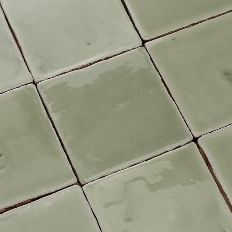 Pale green square ceramic tiles with an irregular glazed surface; dark joints and visible grout, arranged in a mosaic pattern with slightly misaligned edges.