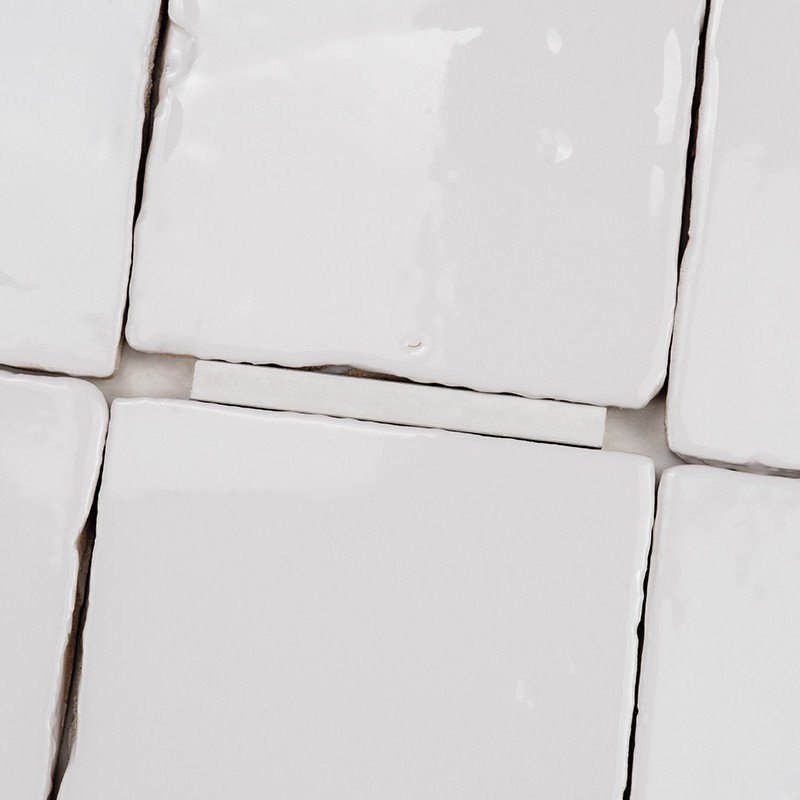 Azulejos blancos esmaltados, cuadrados y ligeramente irregulares; juntas visibles con lechada gris y pequeñas hendiduras; vista en primer plano de varios azulejos contiguos.