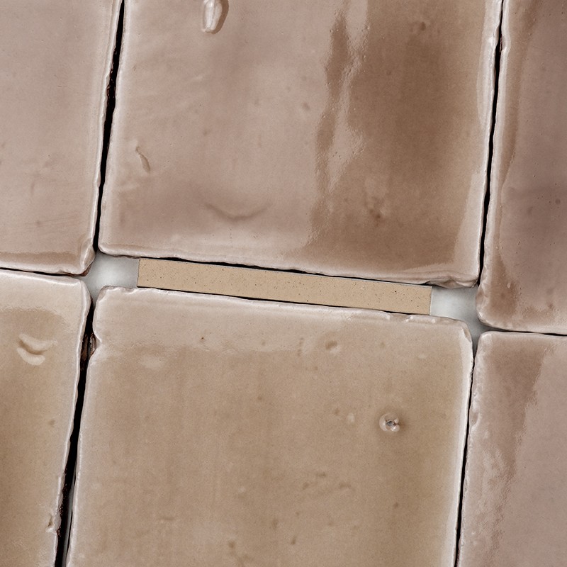 Azulejo beige esmaltado en primer plano; juntas claras y ligeras gotas de agua; bordes irregulares y azulejos contiguos similares formando una rejilla.