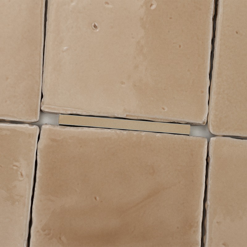 Azulejo cerámico cuadrado color beige con brillo irregular; rodeado por otros azulejos similares y juntas blancas, uno ligeramente desplazado dejando un estrecho hueco.