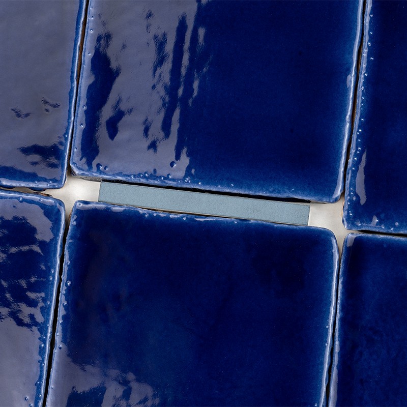 Intense blue square ceramic tiles with glossy glaze, white grout between them; grid arrangement, reflective surface and slightly irregular edges.