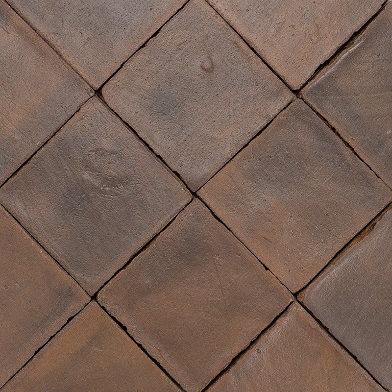 Terracotta tiles arranged in a diamond pattern, rough and worn surface, narrow and dark joints forming a repeated pattern, aged appearance of the pavement.