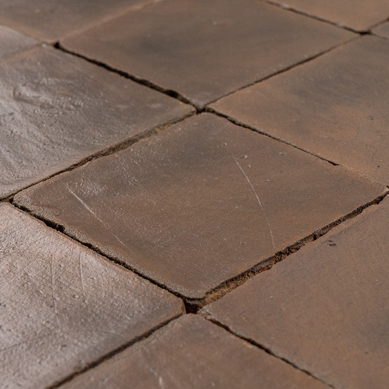 Carrelage de carreaux de terre cuite brun posé au sol, surfaces mates et usées, joints larges et irréguliers, arêtes arrondies et petites fissures visibles.
