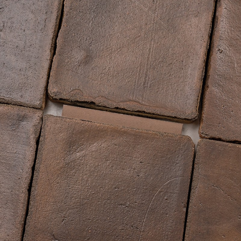 Carrelage en carreaux carrés de terre cuite brun, surfaces mates et usées; joints blancs visibles, bords ébréchés et surfaces légèrement irrégulières en gros plan.