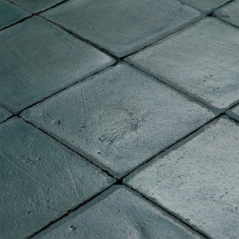 Azulejos cuadrados verde oscuro, superficie rugosa y ligeramente desgastada; juntas estrechas que forman un suelo continuo en perspectiva.