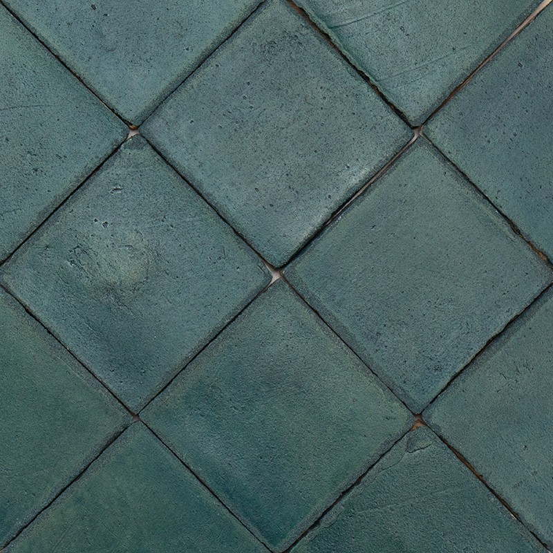 Square tiles arranged in a rhombus, bluish-green color, slightly rough and worn surface; dark joints and small irregularities that create a repetitive pattern.