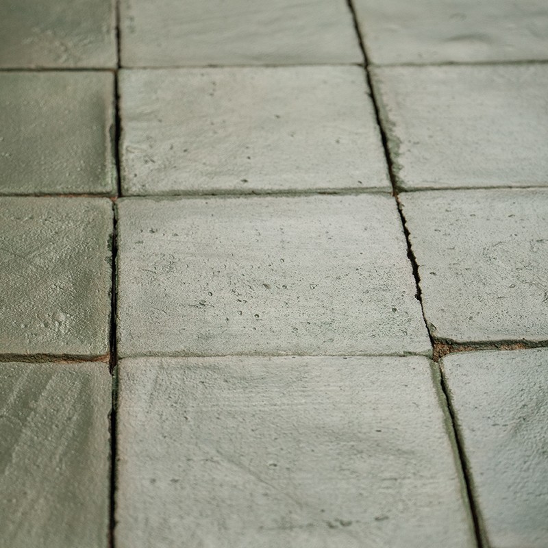 Carrelage de carreaux carrés vert-gris usés, joints étroits et fissurés, surface légèrement brillante; plan rapproché montrant alignement imparfait et traces d'usure.
