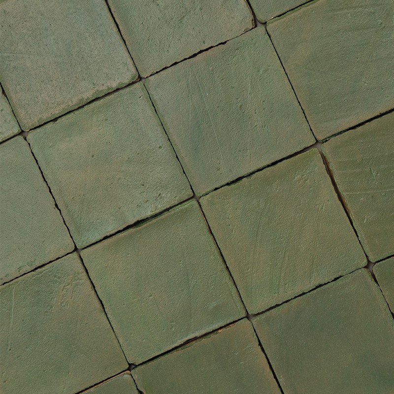 Carrelage vert mat de carreaux carrés légèrement irréguliers, joints sombres formant un quadrillage; surface texturée et usée, disposition légèrement décalée en diagonale.