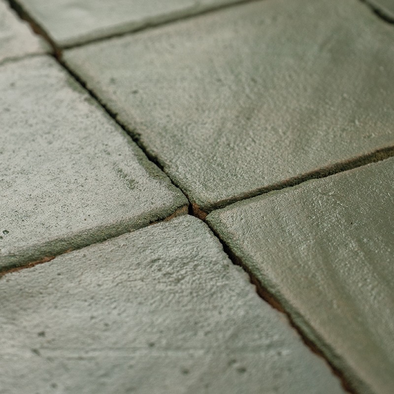 Carrelage en carreaux carrés vert-gris, surface légèrement texturée et usée; joints étroits foncés formant une croix au centre, éclairage doux révélant reflets et irrégularités.